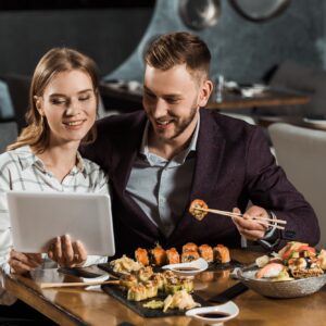 Attractive couple of yound adult using digital tablet while eating sushi in restaurant