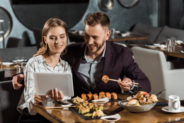 Attractive couple of yound adult using digital tablet while eating sushi in restaurant